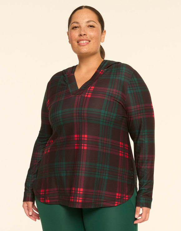 A woman wearing a red and green plaid hooded top and green leggings stands against a plain, light-colored background.