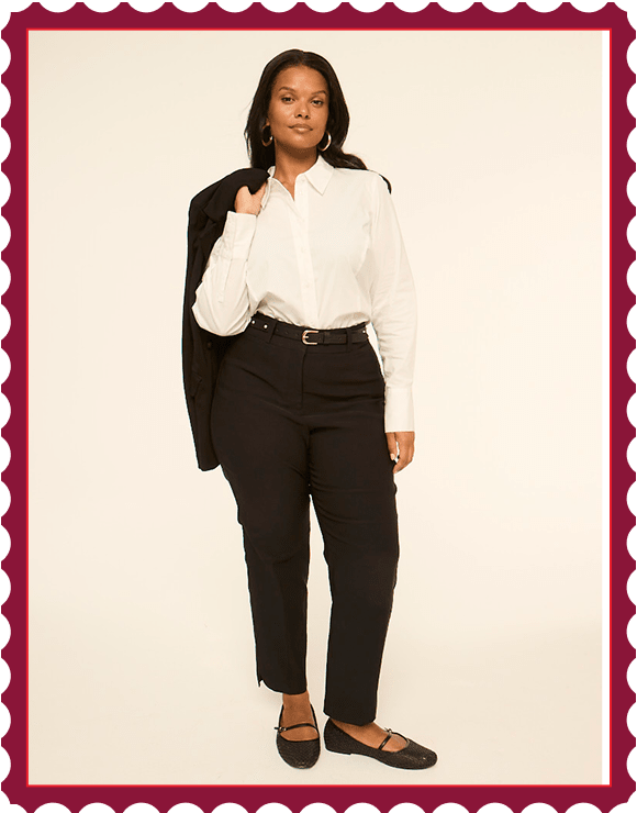 A woman in a white blouse and black pants stands against a plain, light-colored background, with a black blazer draped over her shoulder.