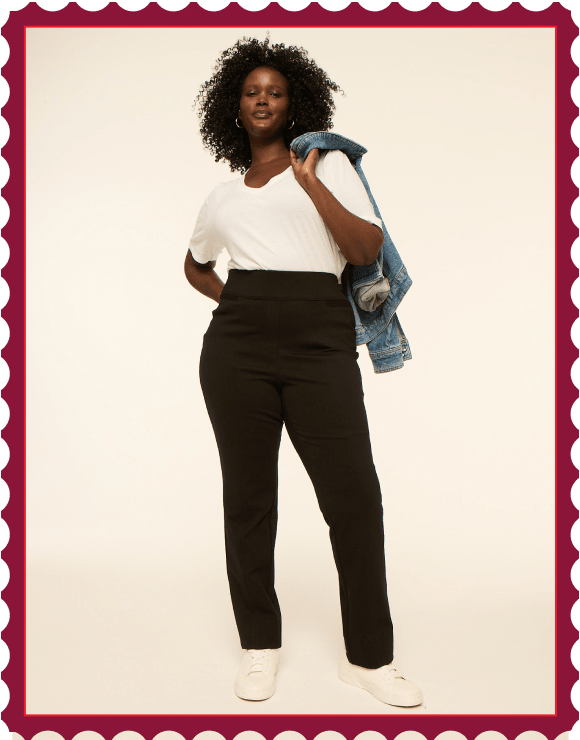 A woman in a white t-shirt, black pants, and white sneakers stands with a denim jacket slung over her shoulder against a plain, light-colored background.