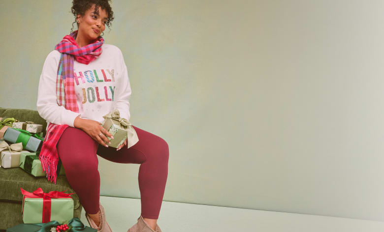 A woman wearing a festive 'Holly Jolly' sweater and colorful scarf sits among wrapped gifts against a plain, light-colored background.