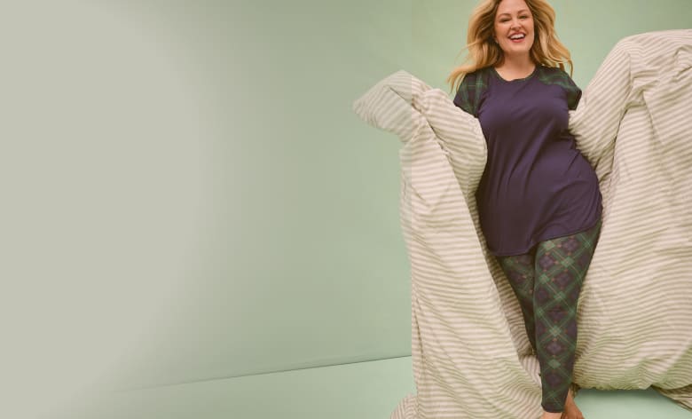A smiling woman in navy and plaid pajamas stands wrapped in a white comforter against a plain light green background.