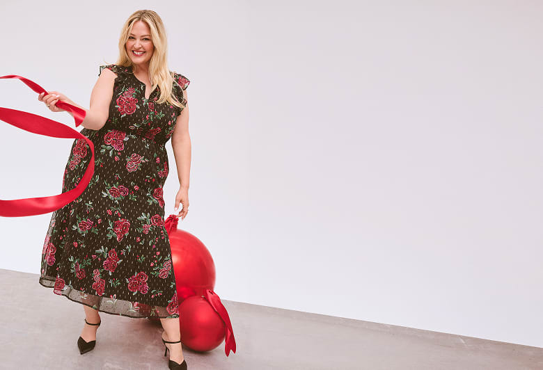 A smiling woman in a flowery black dress holding swinging a red scarf stands wrapped in front of a light background with a large red christmas ornament.