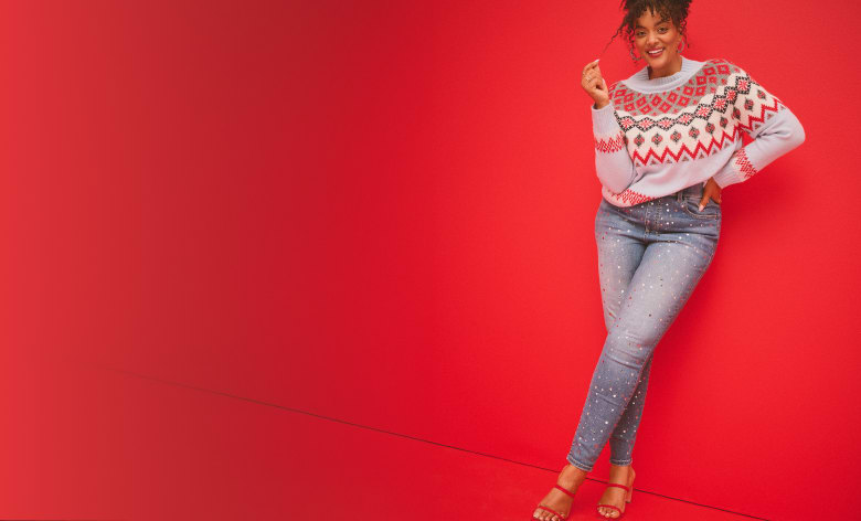 A woman wearing a festive sweater and jeans stands against a bright red background.