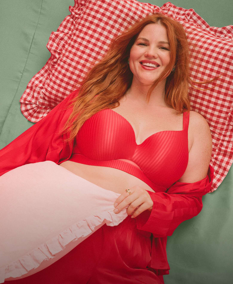 A woman with red hair wearing a red lingerie set poses against a backdrop of draped red satin fabric.