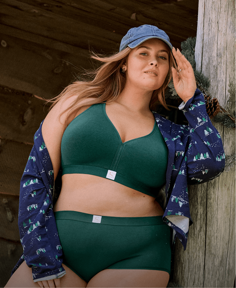 A woman wearing red and green plaid lingerie and an open matching shirt poses in front of a rustic stone fireplace decorated with pine branches and pinecones.