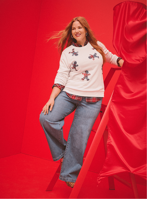 A woman wearing a green 'Frosty' sweater and plaid pants leans against a red North Pole mailbox against a plain light-blue background.