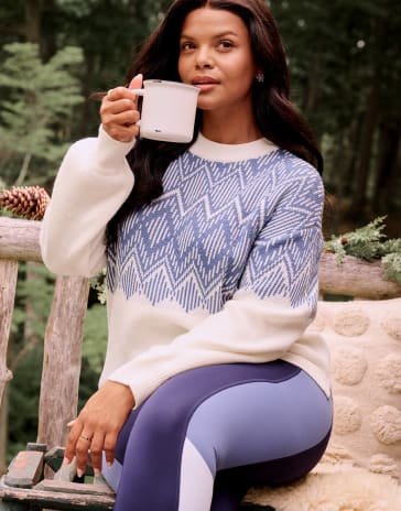 A woman in a pink longsleeve tshirt and black pants stands in front of a cream colored background with a black longsleeve shirt around her neck.