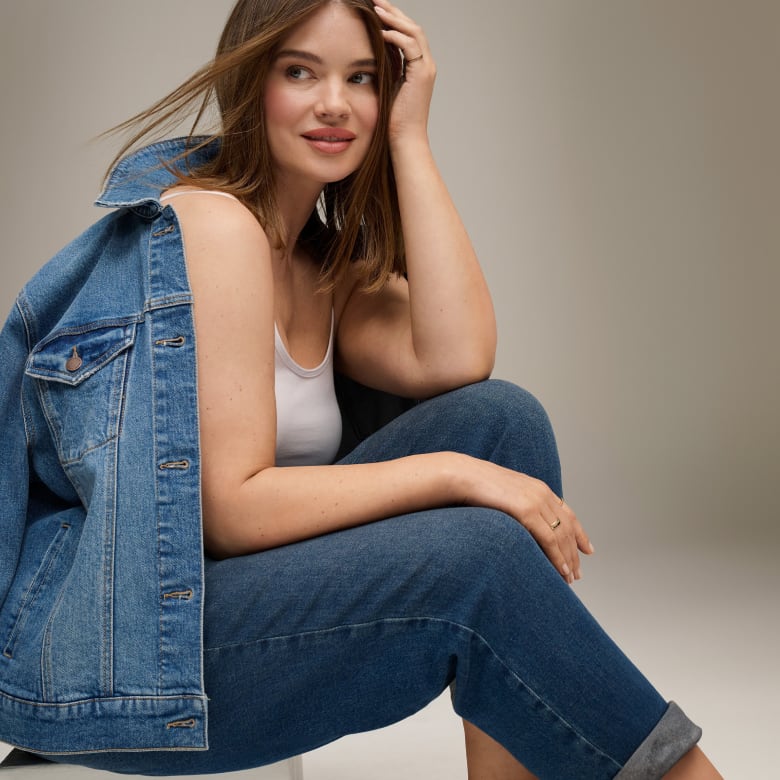 A woman in a white tank top and jeans, with a denim jacket draped over her shoulder, poses against a plain, light gray background.