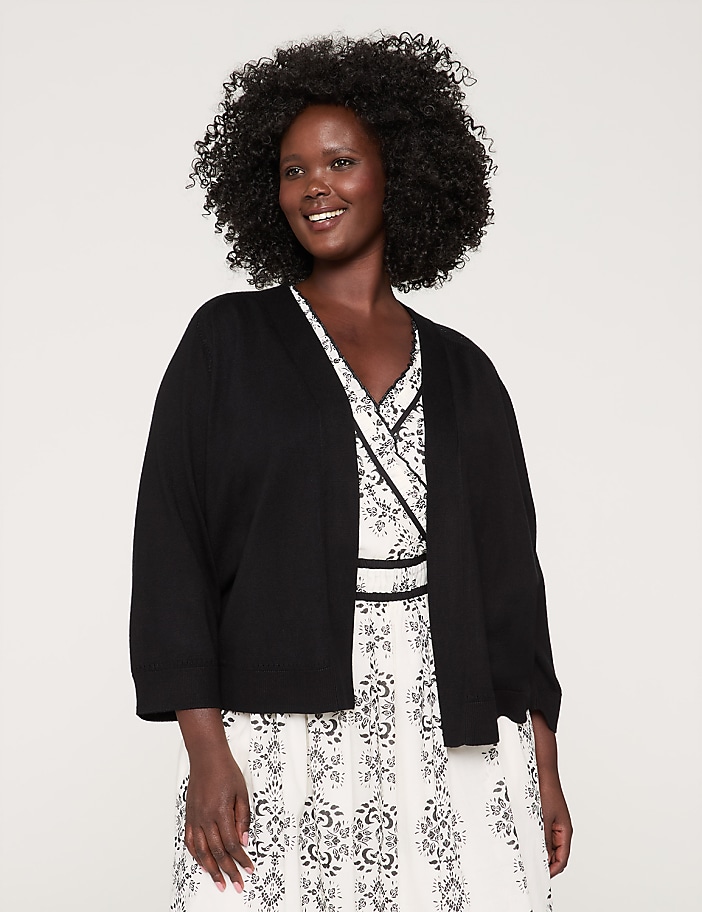 A smiling woman with curly dark hair wearing a black jacket and a white patterned dress stands against a plain background.