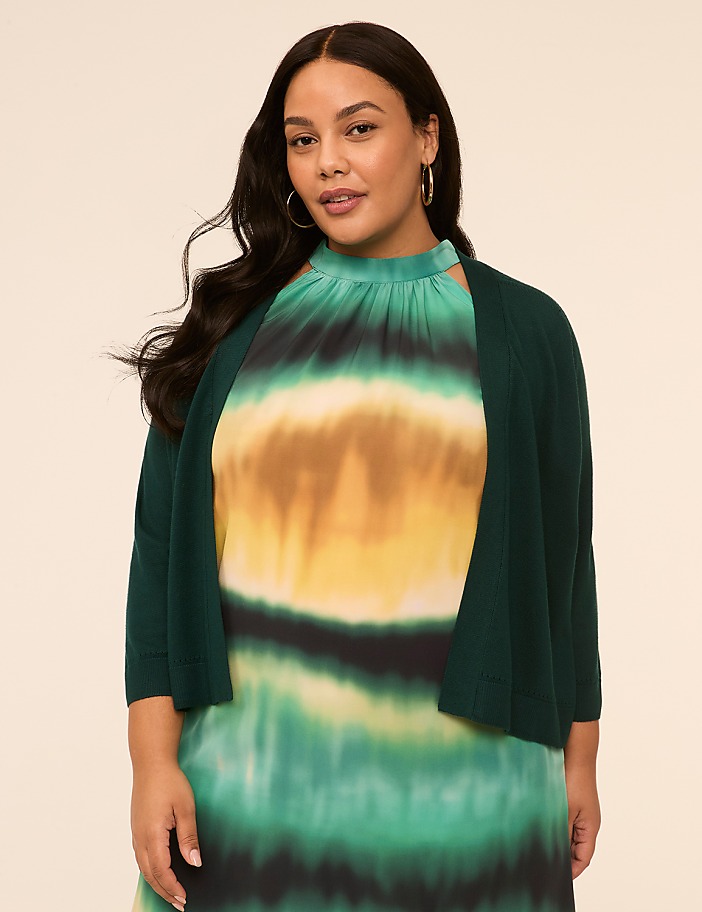 A young woman with long dark hair is wearing a vibrant green, yellow, and black patterned dress and standing against a plain beige background.