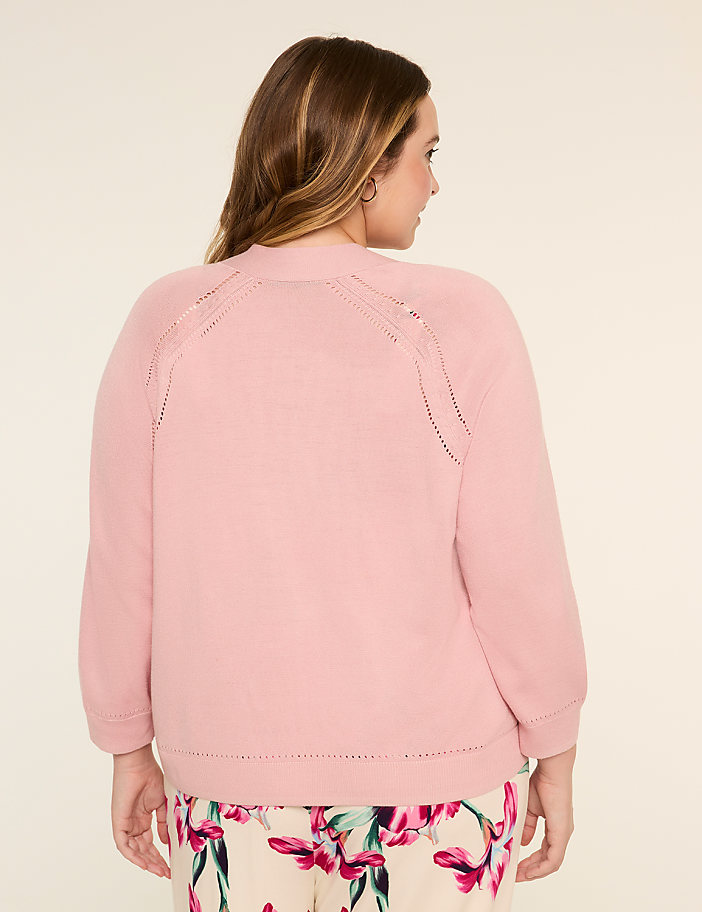 A woman with long, wavy brown hair wearing a pink sweater stands against a plain, light-colored background.