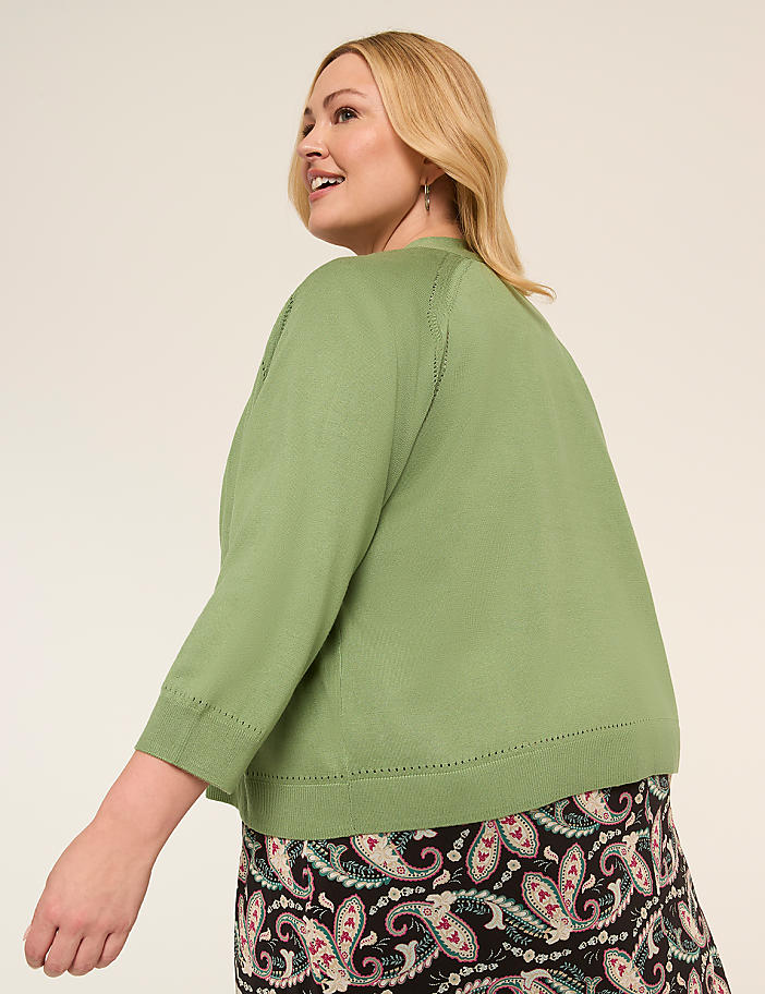 A smiling woman with blonde hair wearing a green top and a patterned skirt stands against a plain background.