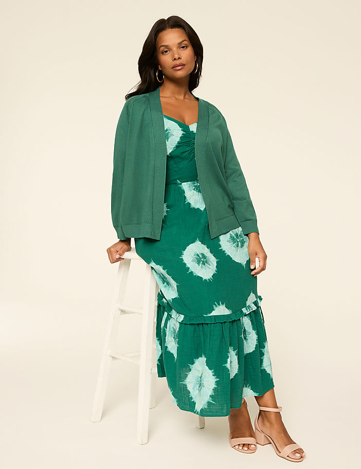 The image shows a woman wearing a green and white patterned dress and a green jacket, standing against a plain light-colored background.