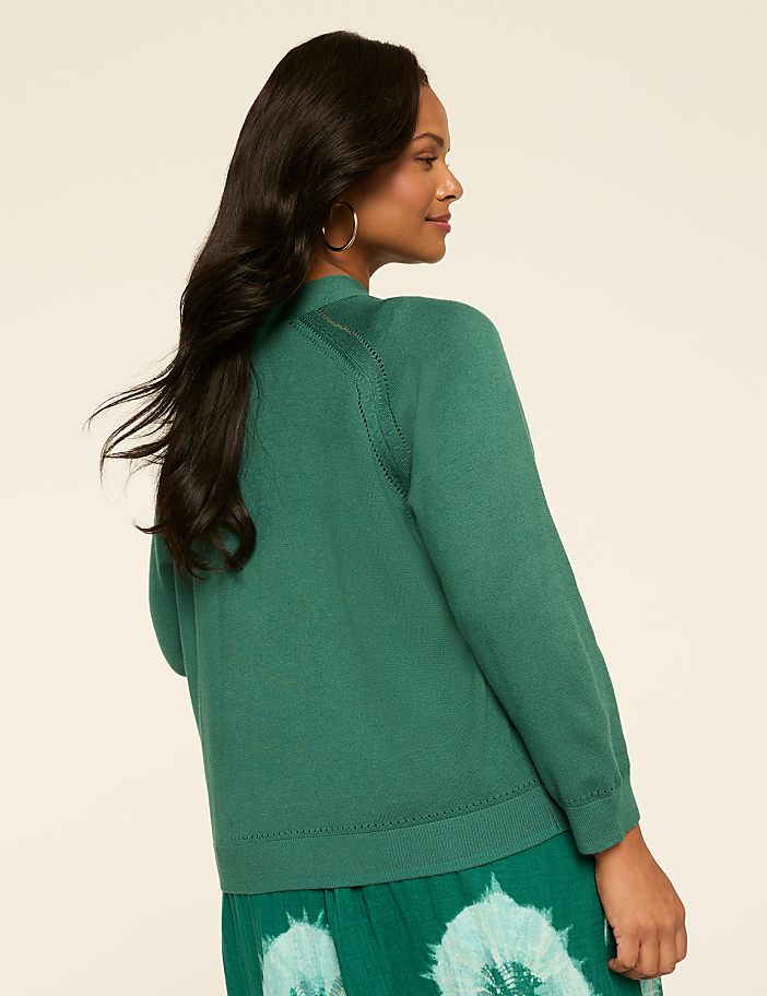 A woman with long, dark hair wearing a green sweater stands against a plain, light-colored background.