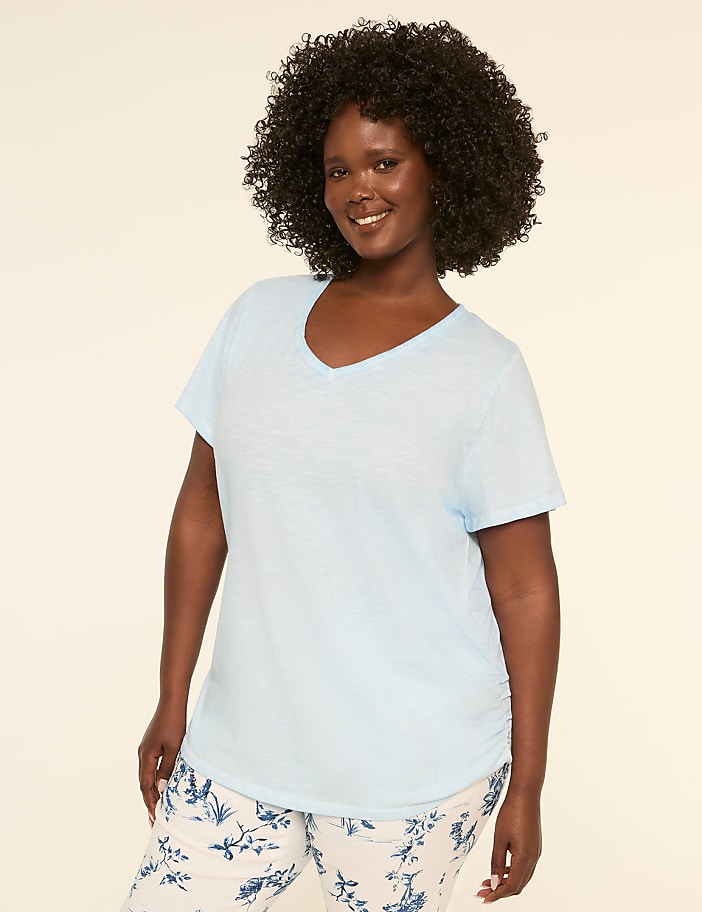 A smiling woman with curly dark hair wearing a light blue top stands against a plain beige background.