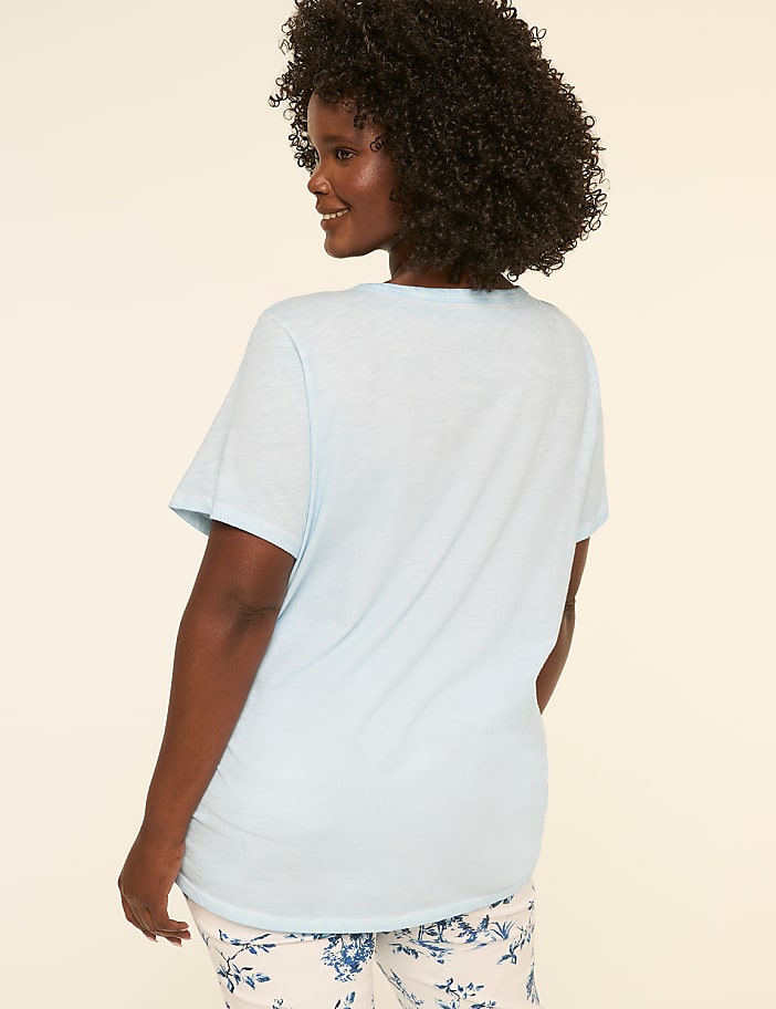 A person with curly dark hair wearing a white t-shirt stands against a plain light-colored background.