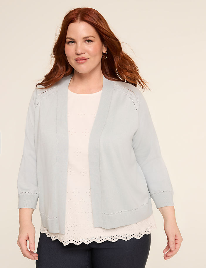A smiling woman with long, wavy red hair wearing a white cardigan and a white top stands against a plain background.