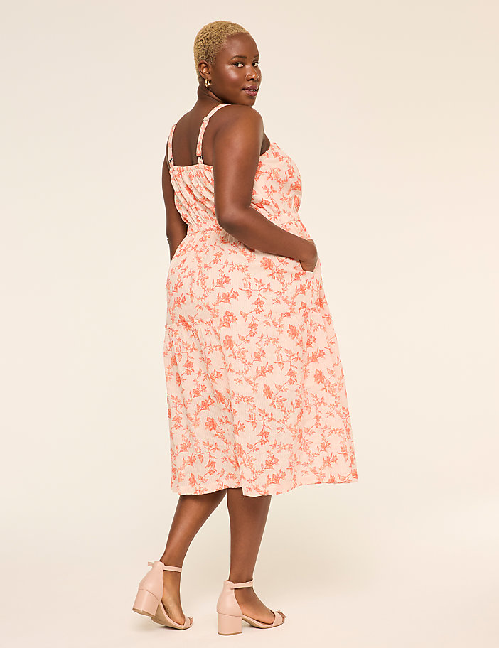 A woman wearing a pink floral dress stands against a plain beige background, her hair styled in a short, natural cut.