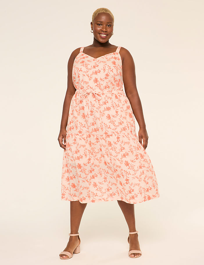 A smiling person wearing a pink floral dress stands against a plain beige background.