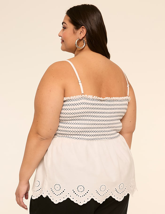 A woman with long dark hair wearing a white sleeveless top with a scalloped hem stands against a plain beige background.
