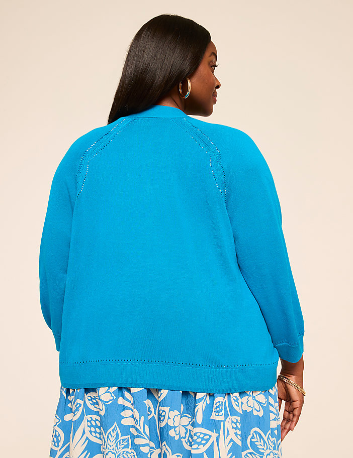 A woman wearing a bright blue sweater stands against a plain beige background, her long dark hair visible from behind.