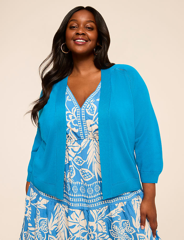 A smiling woman with long dark hair wearing a bright blue top and a patterned dress stands against a plain beige background.