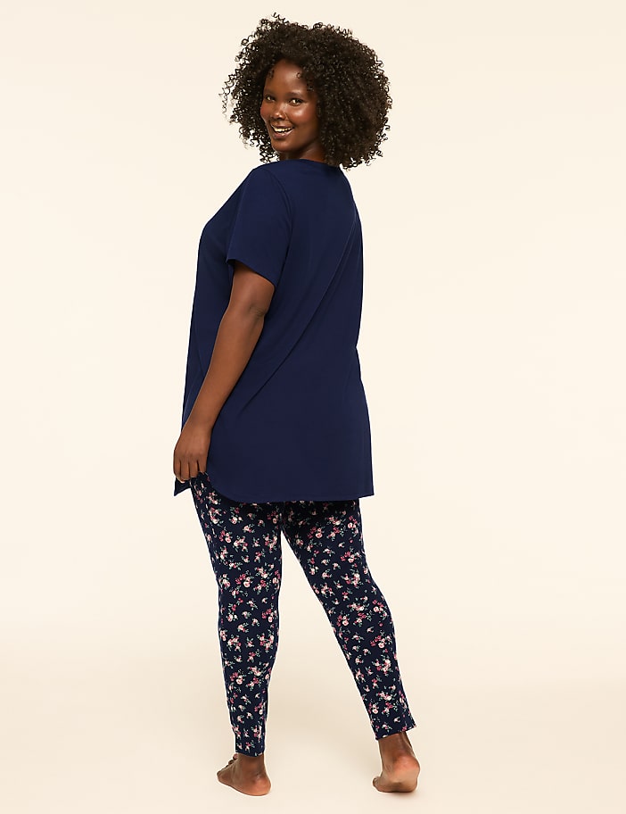 A woman with dark curly hair smiles over her shoulder while wearing a navy blue t-shirt and floral leggings against a plain cream background.