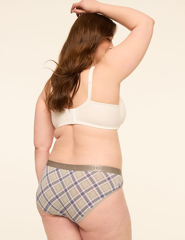 A woman in a white bra and plaid underwear stands with her arm raised against a plain, light-colored background.