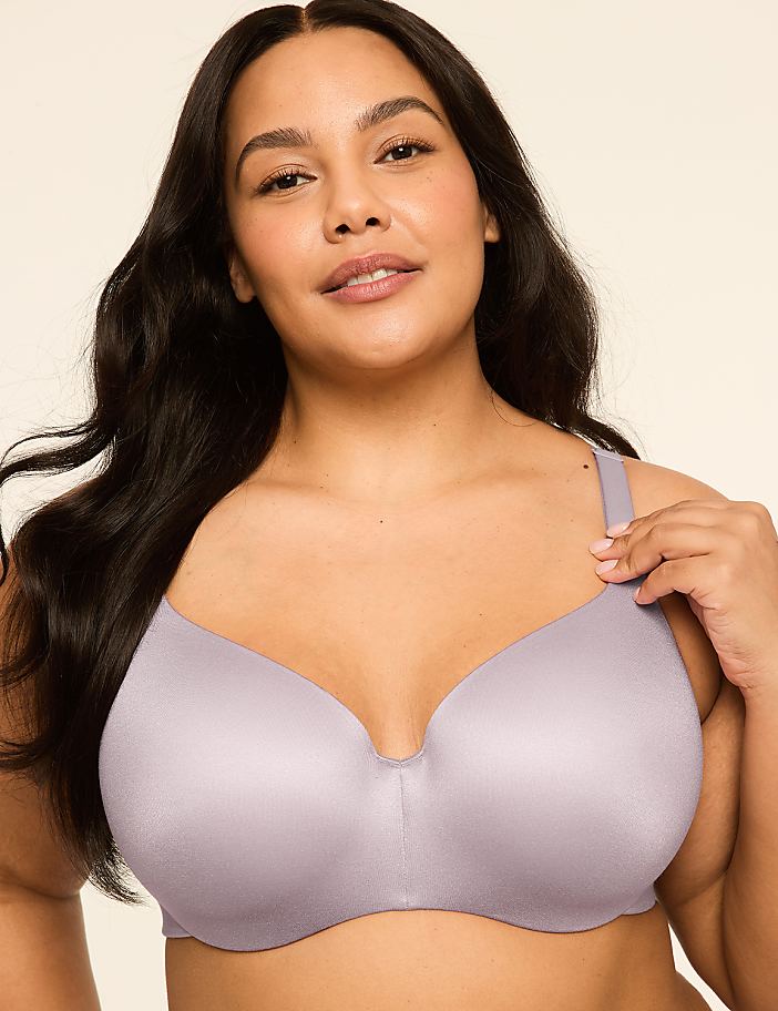 A woman with dark hair and a light complexion wears a light purple bra, with a plain, light-colored background.
