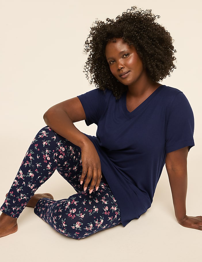 A woman with dark curly hair sits on a plain background wearing a navy blue v-neck shirt and floral patterned leggings.