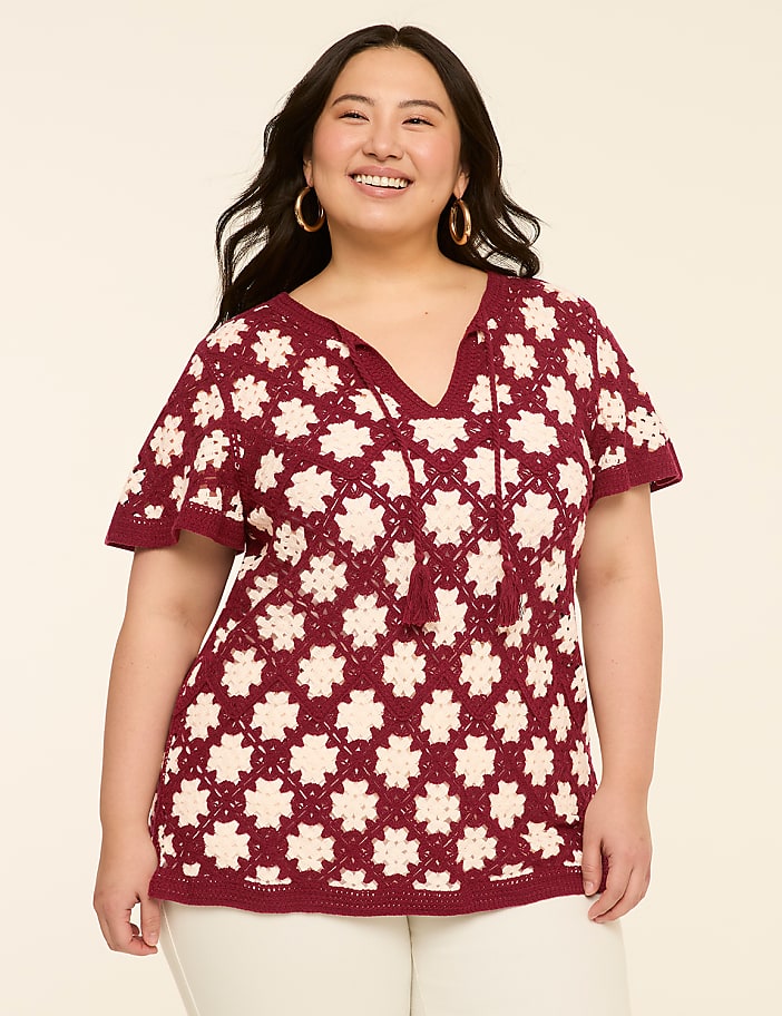 A smiling woman in a maroon and cream crocheted top stands against a plain, light beige background.
