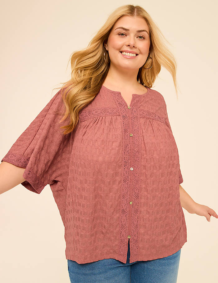 A smiling woman with blonde hair and a pink blouse stands against a plain, light-colored background.