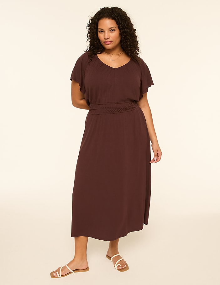 A woman in a dark brown dress and white sandals stands against a plain, light-colored background.