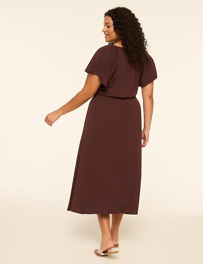 A woman in a dark brown dress stands against a plain, light brown background.