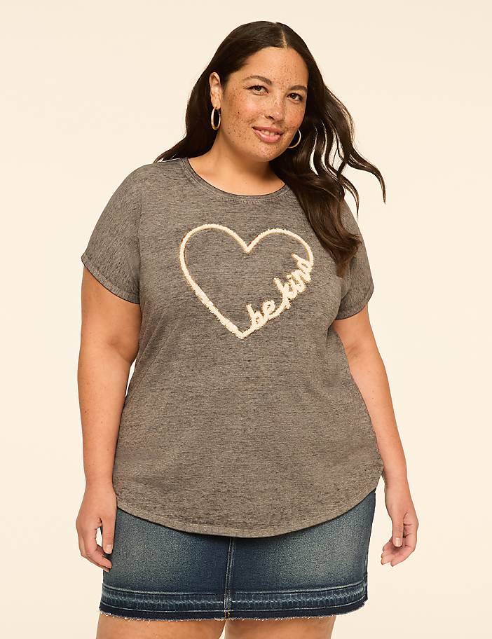 A woman in a grey t-shirt with a heart design and a denim skirt stands against a plain, light-colored background.