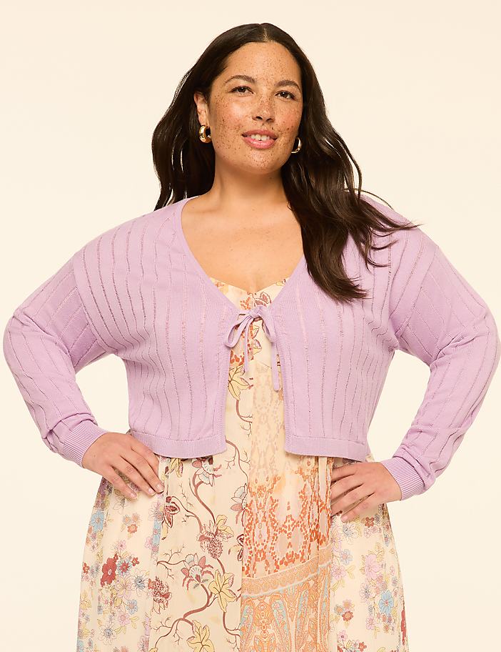 A woman in a floral dress and a lavender cardigan stands against a plain, light-colored background.