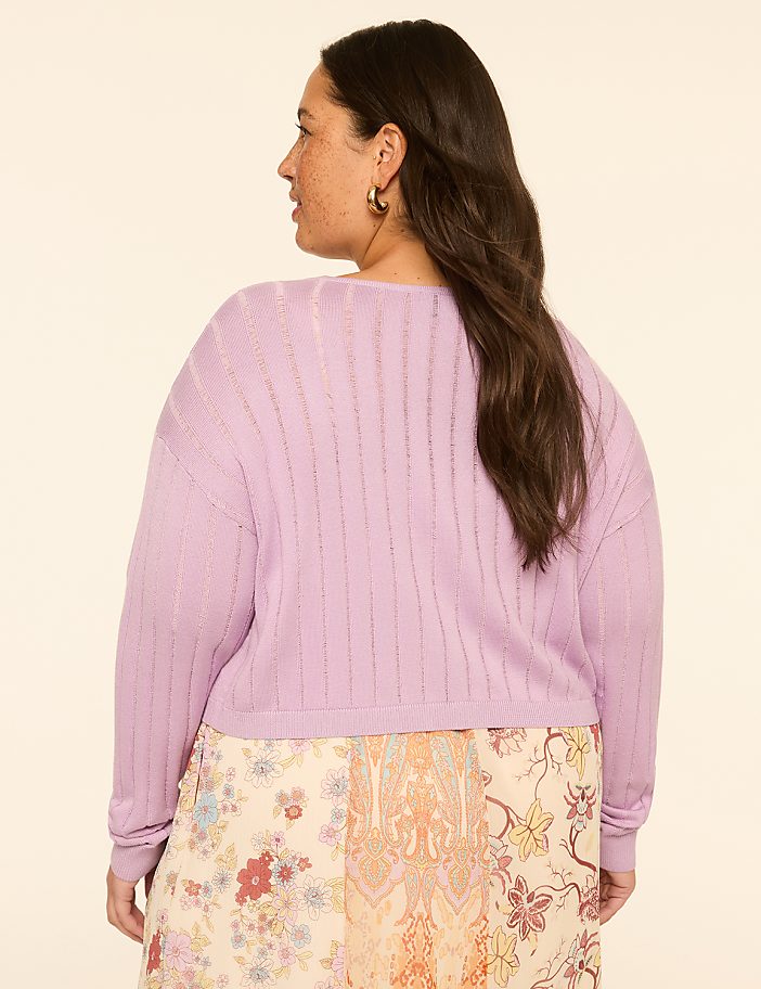 A woman in a light purple sweater and a floral skirt stands against a plain, light-colored background.