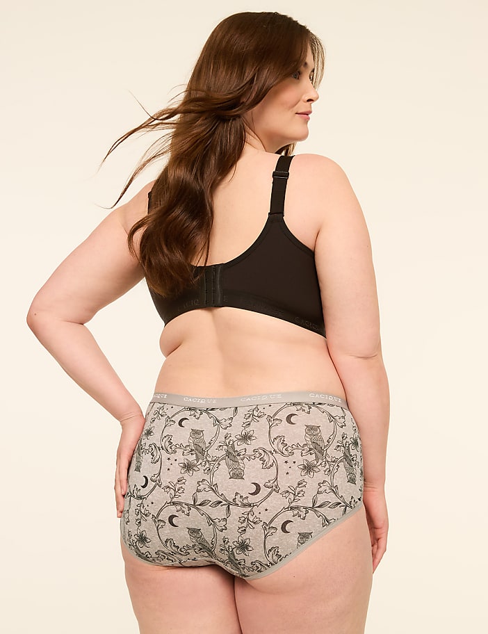 A woman with long brown hair wears a black bra and grey patterned underwear with owls and moons, against a plain cream background.