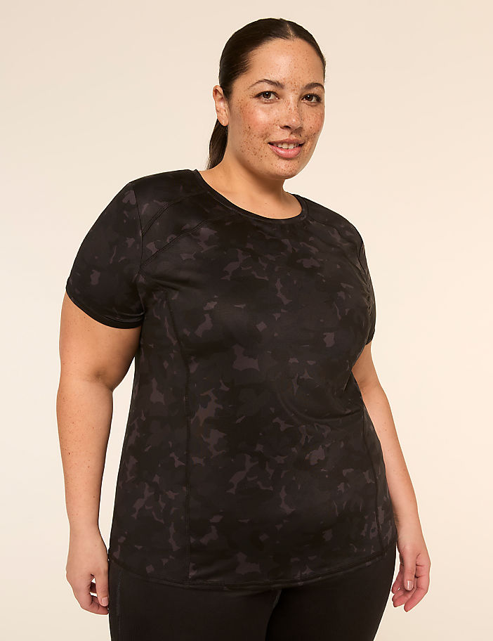 A woman with dark hair pulled back stands in the foreground wearing a black patterned t-shirt and black leggings, against a plain, light-colored background.