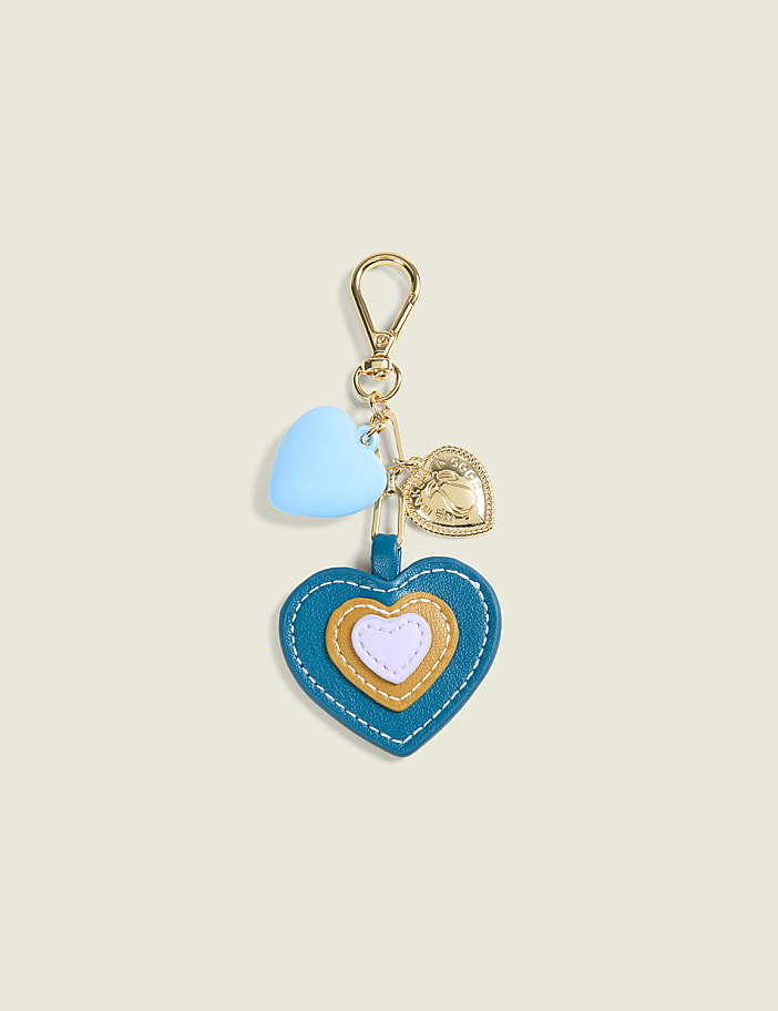 A teal, heart-shaped keychain with layered hearts and small charms hangs against a pale yellow background.