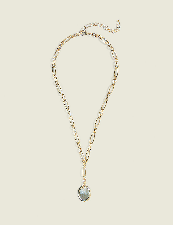 A gold necklace with an oval pendant hangs against a plain, light beige background.