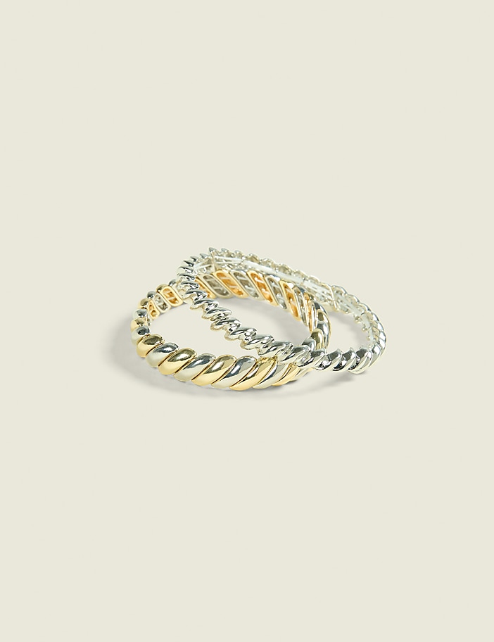 Two twisted rings, one with gold and silver sections and the other entirely silver, rest on a plain, light-colored background.