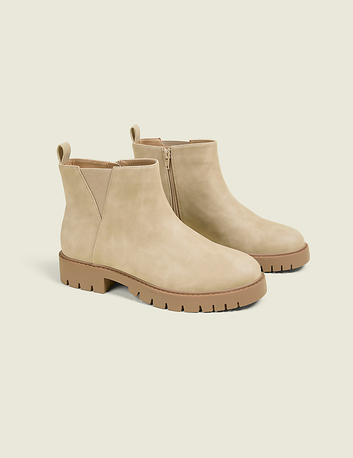 A pair of beige suede Chelsea boots with thick tan soles are displayed against a plain, light beige background.