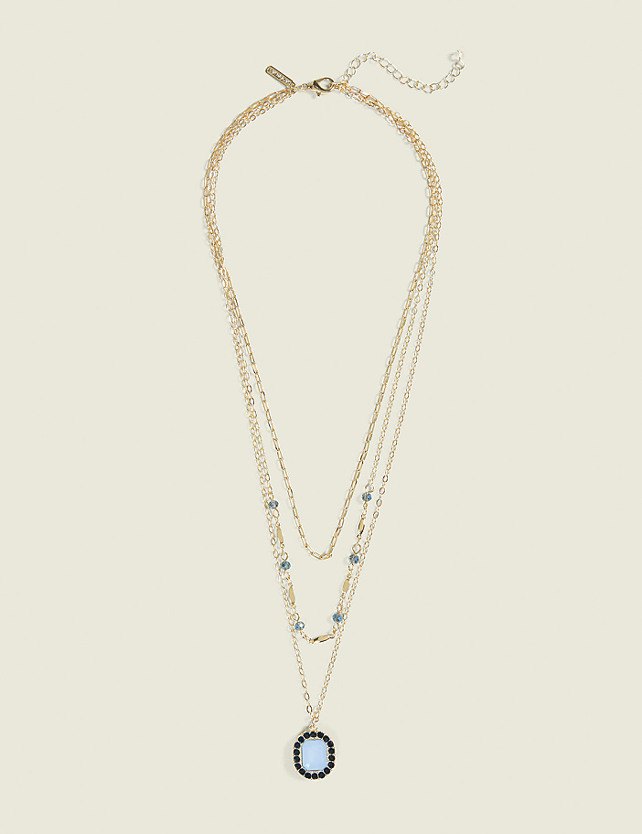 A layered gold necklace with a blue pendant rests against a plain, light beige background.
