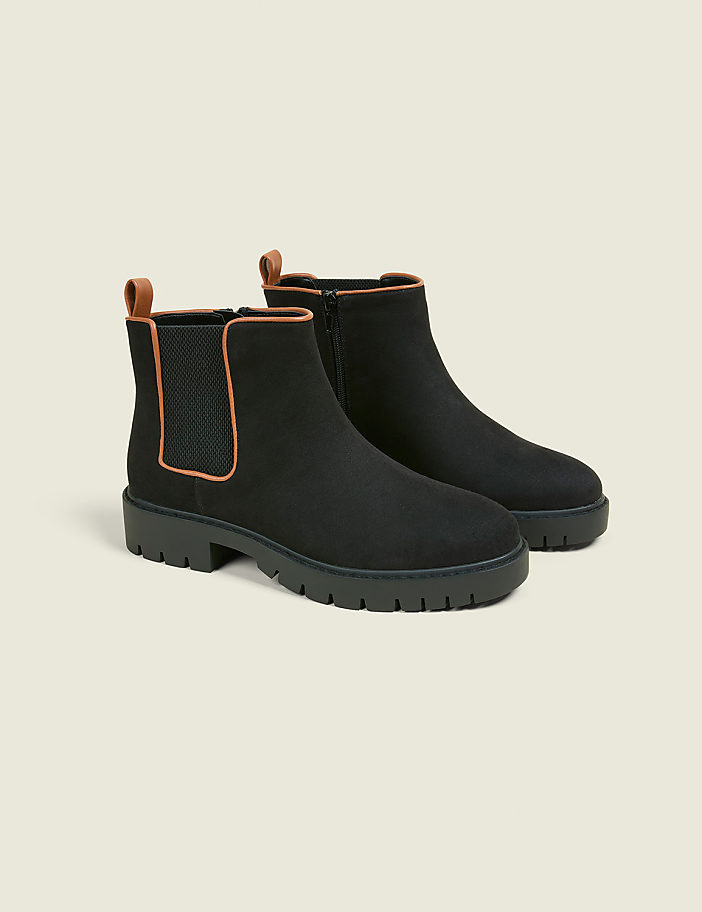 A pair of black Chelsea boots with tan accents and thick soles rests on a light beige background.