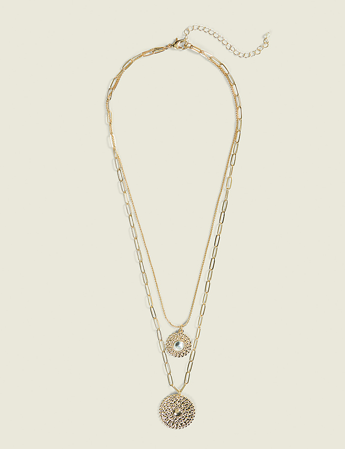 A layered gold necklace with two circular pendants hangs against a plain, light beige background.