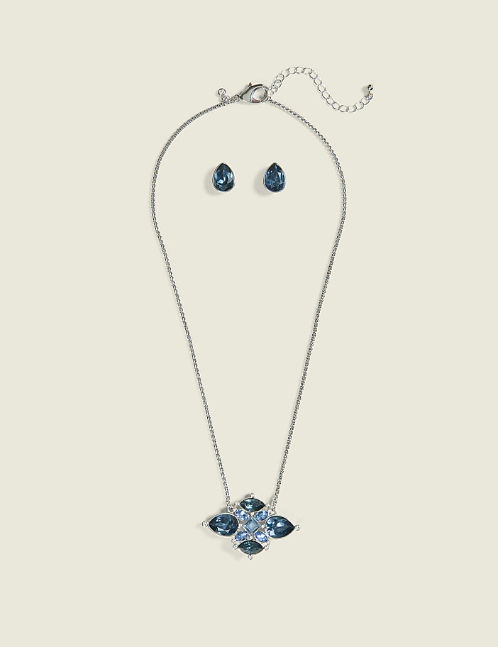 A silver necklace with a blue jeweled pendant and matching teardrop earrings are displayed against a plain, light-colored background.