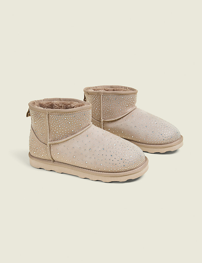A pair of beige, rhinestone-embellished ankle boots with fluffy lining are displayed against a plain, light beige background.