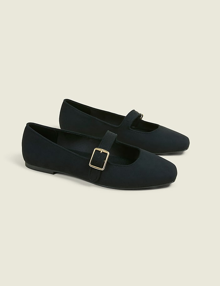 A pair of black Mary Jane flats with gold buckles rests on a light beige background.