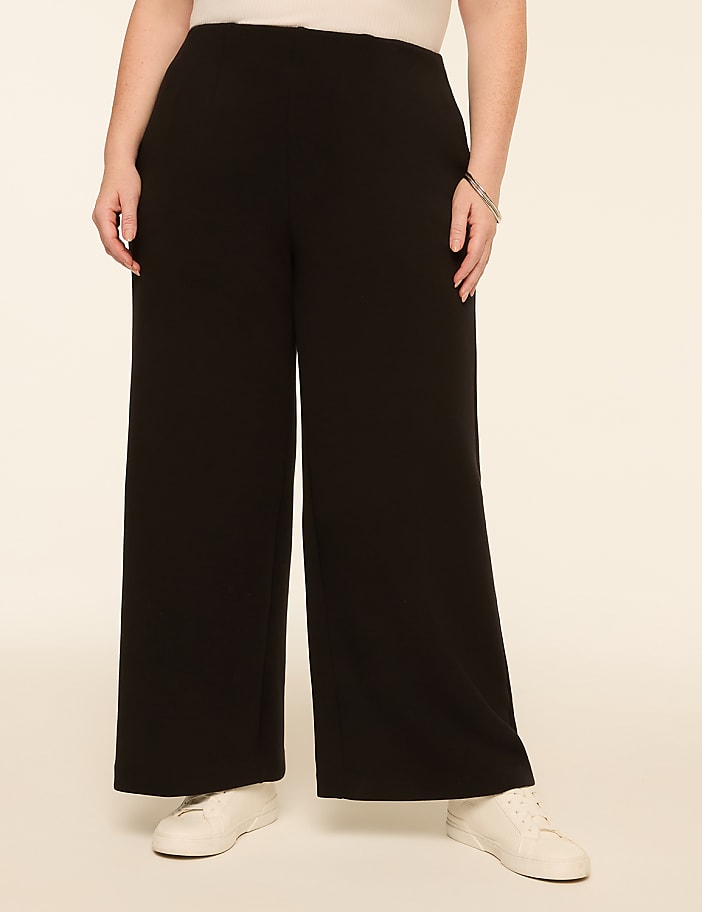 A person wearing black wide-leg pants and white sneakers stands against a plain, light-colored background.